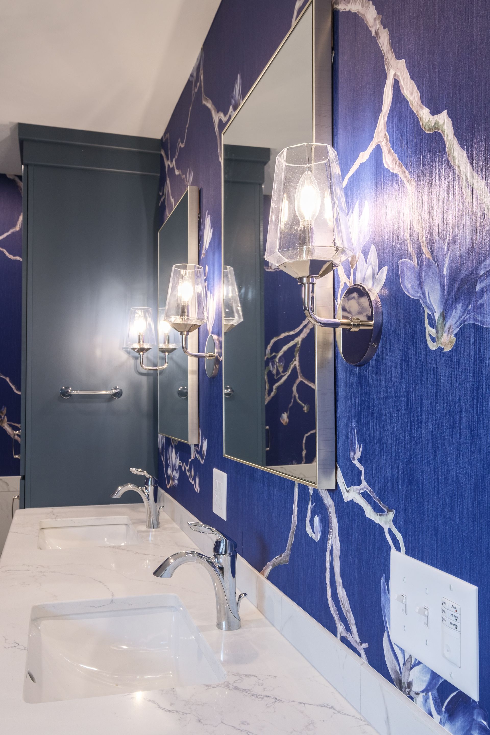 Blue floral wallpapered bathroom with sinks, mirror, and sconces.