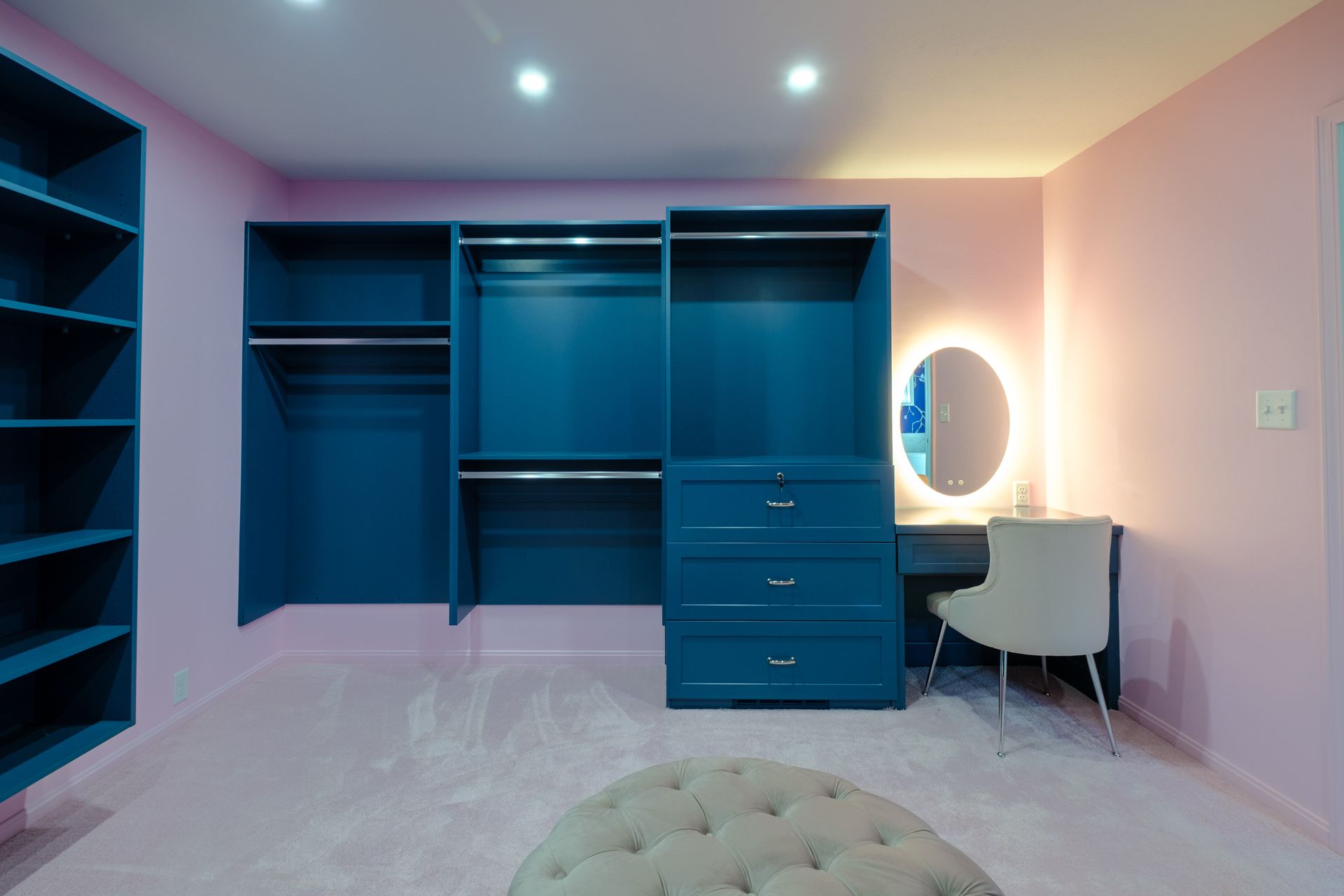 Pink-walled walk-in closet with blue built-ins, vanity, and a tufted ottoman on a light pink carpet.