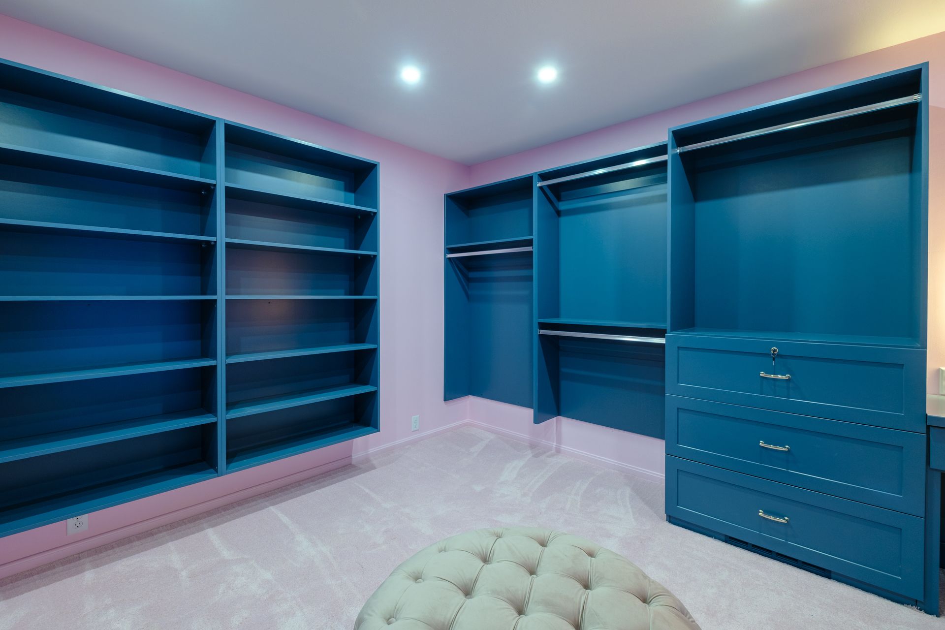 Blue built-in closet with shelves and drawers, set against pink walls and carpet. A cushioned ottoman sits in front.