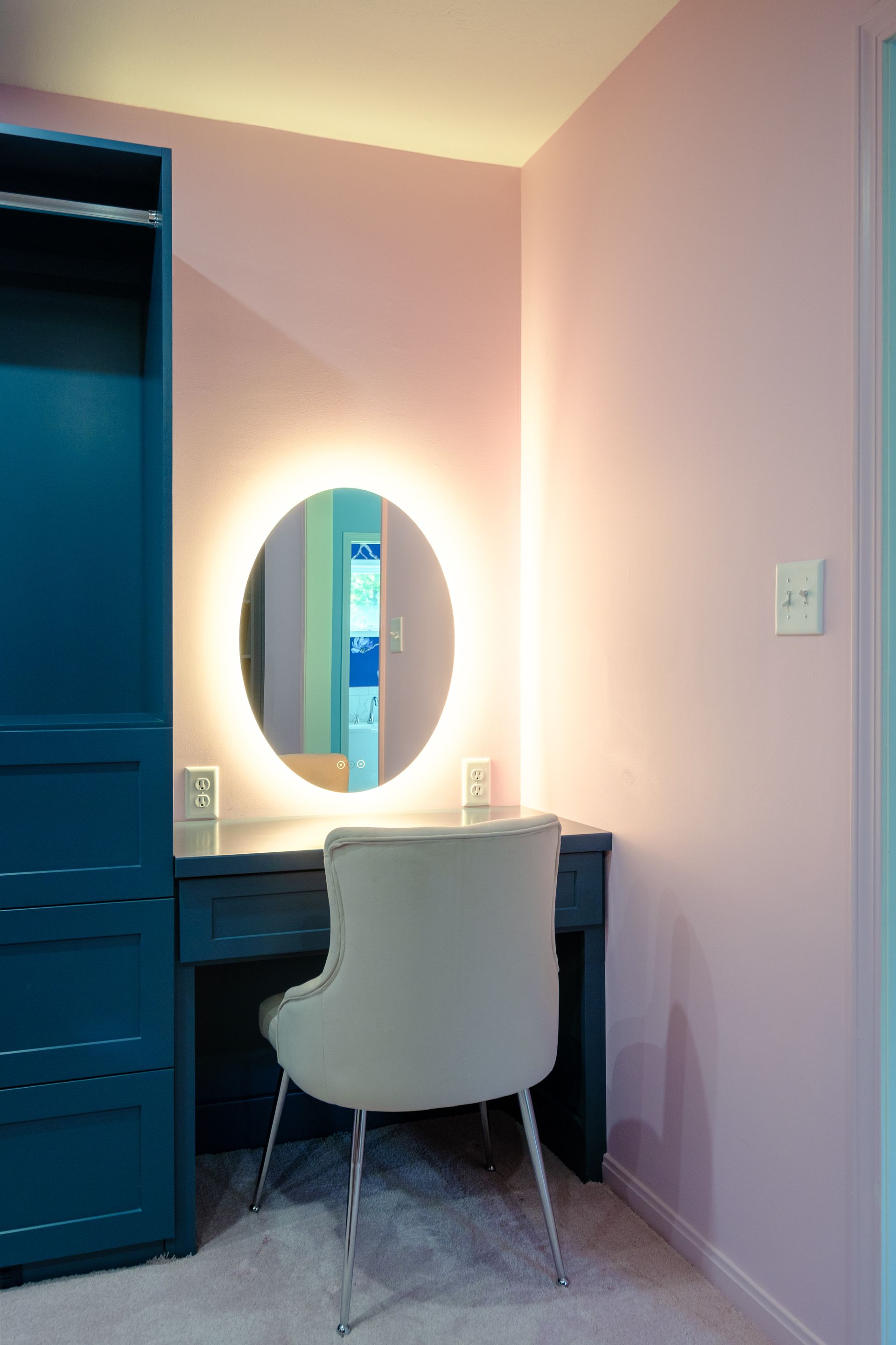 Vanity area with oval lighted mirror, teal vanity and closet, pink walls, and silver-legged chair.