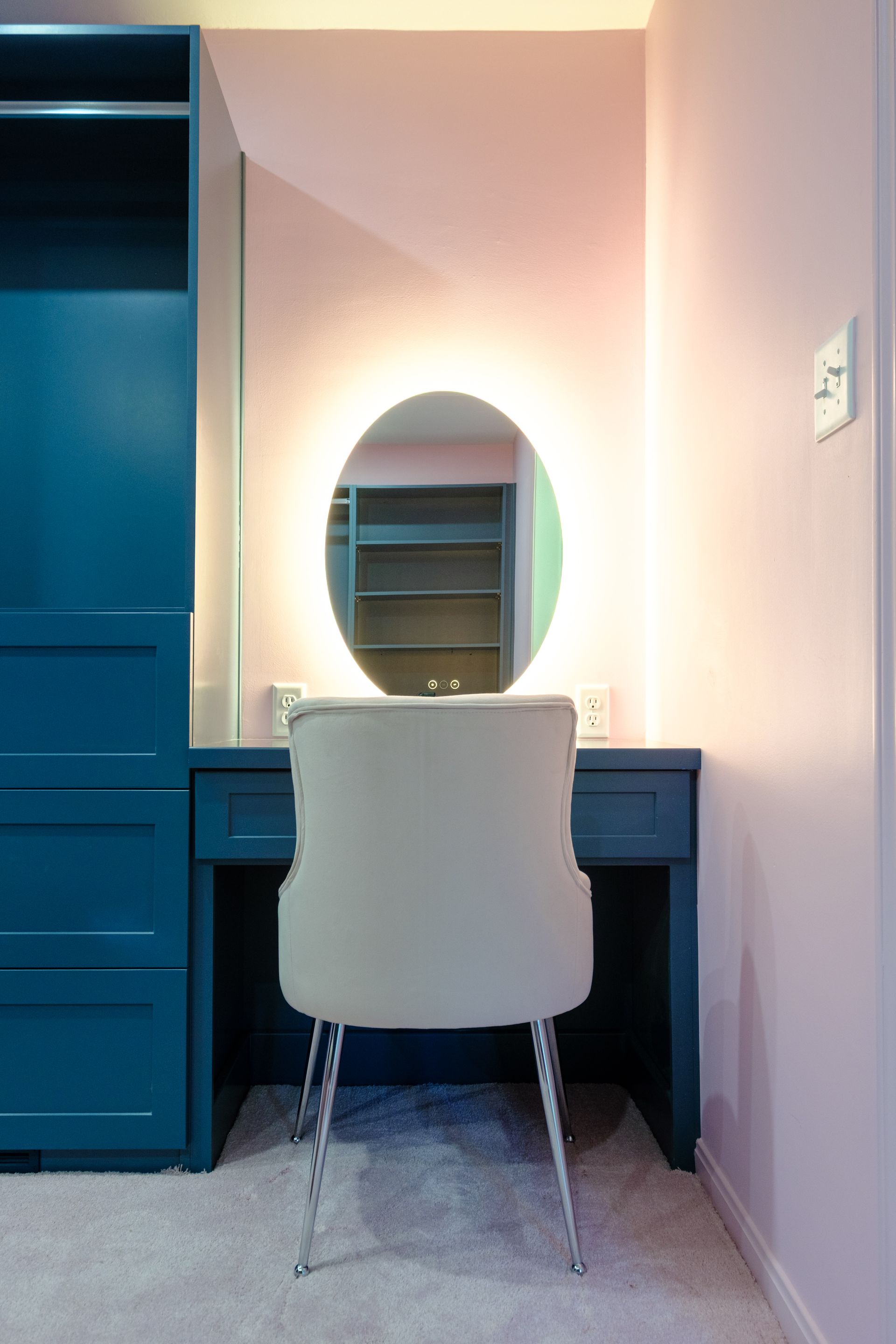 Teal vanity with illuminated oval mirror and white chair against a pink wall. Teal closet on the left.