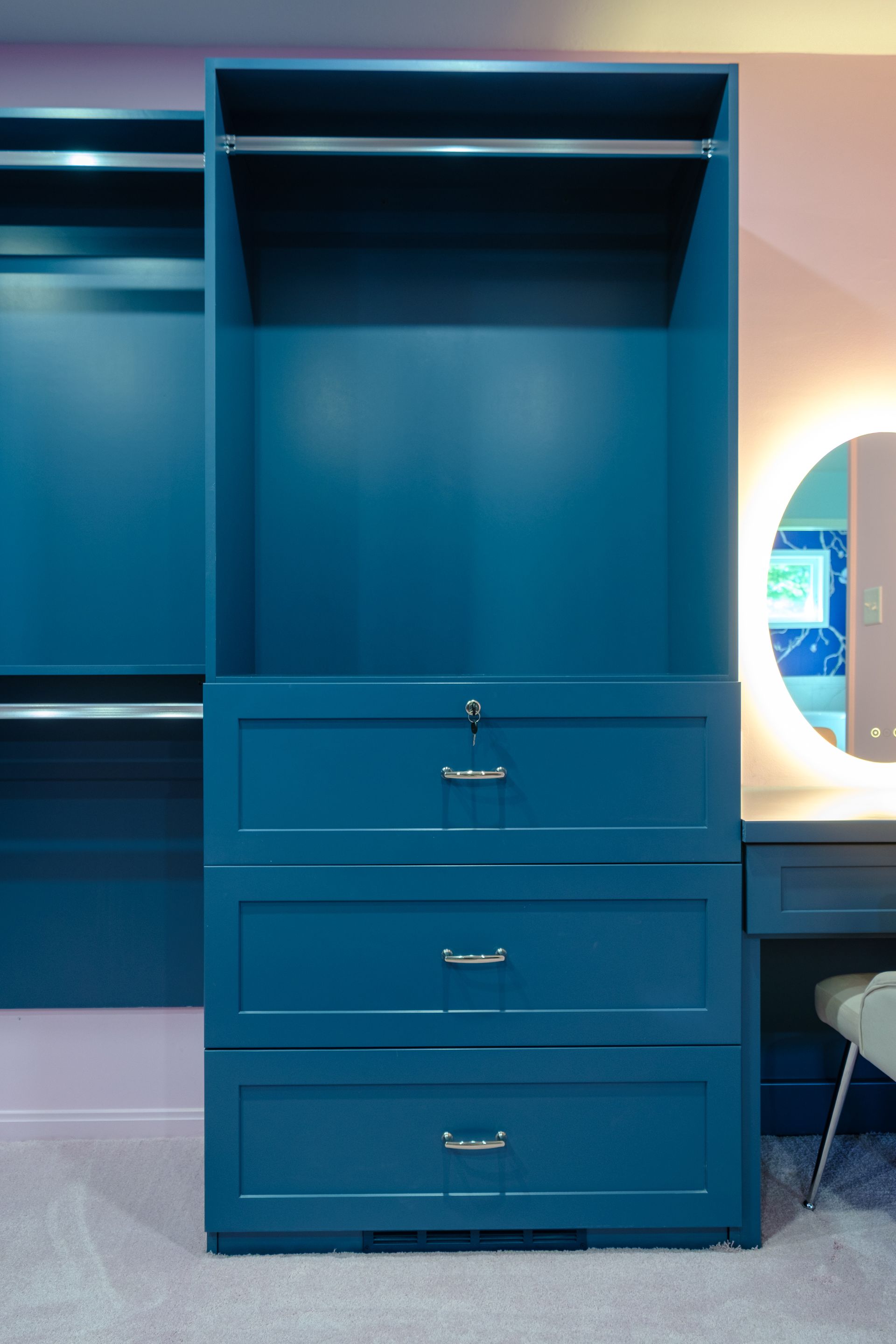 Teal closet with open shelving, drawers, and a rod. A vanity with an illuminated mirror is in the background.