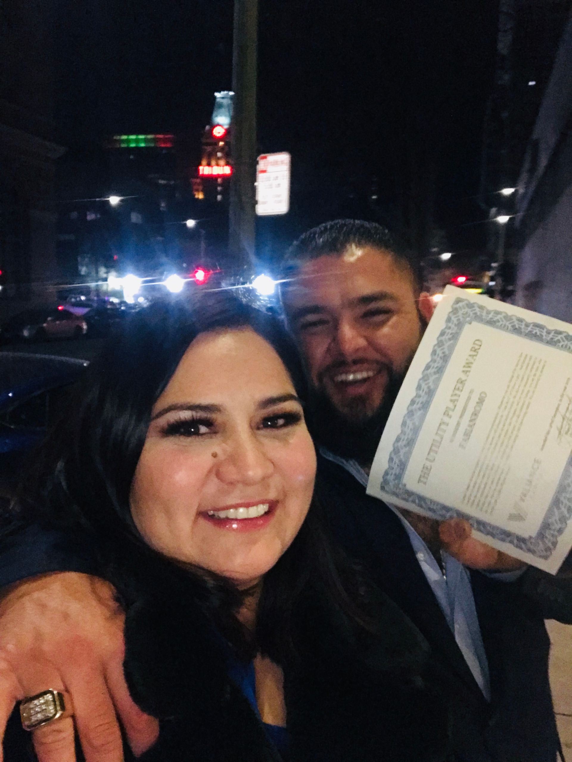 Two smiling people at night; one holds up a document with city lights in the background.