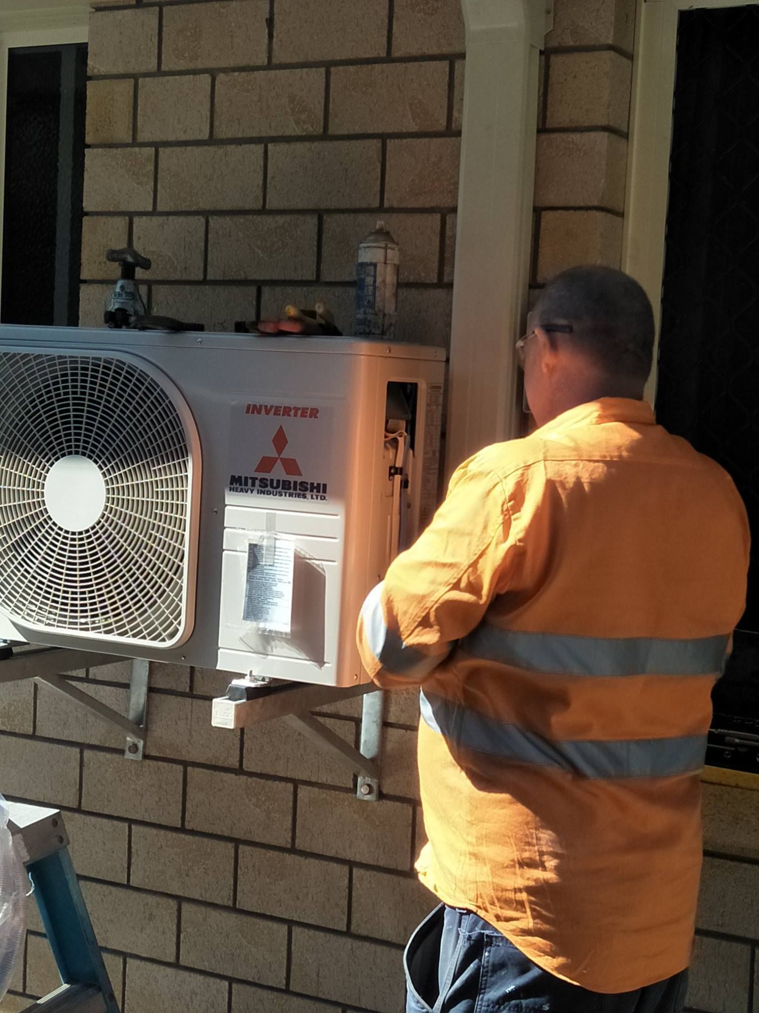 A Man In Orange Work Clothes Working On An Air Conditioning Unit Mounted On A Brick Wall — LSEA Electrical Group in Bucasia, QLD.