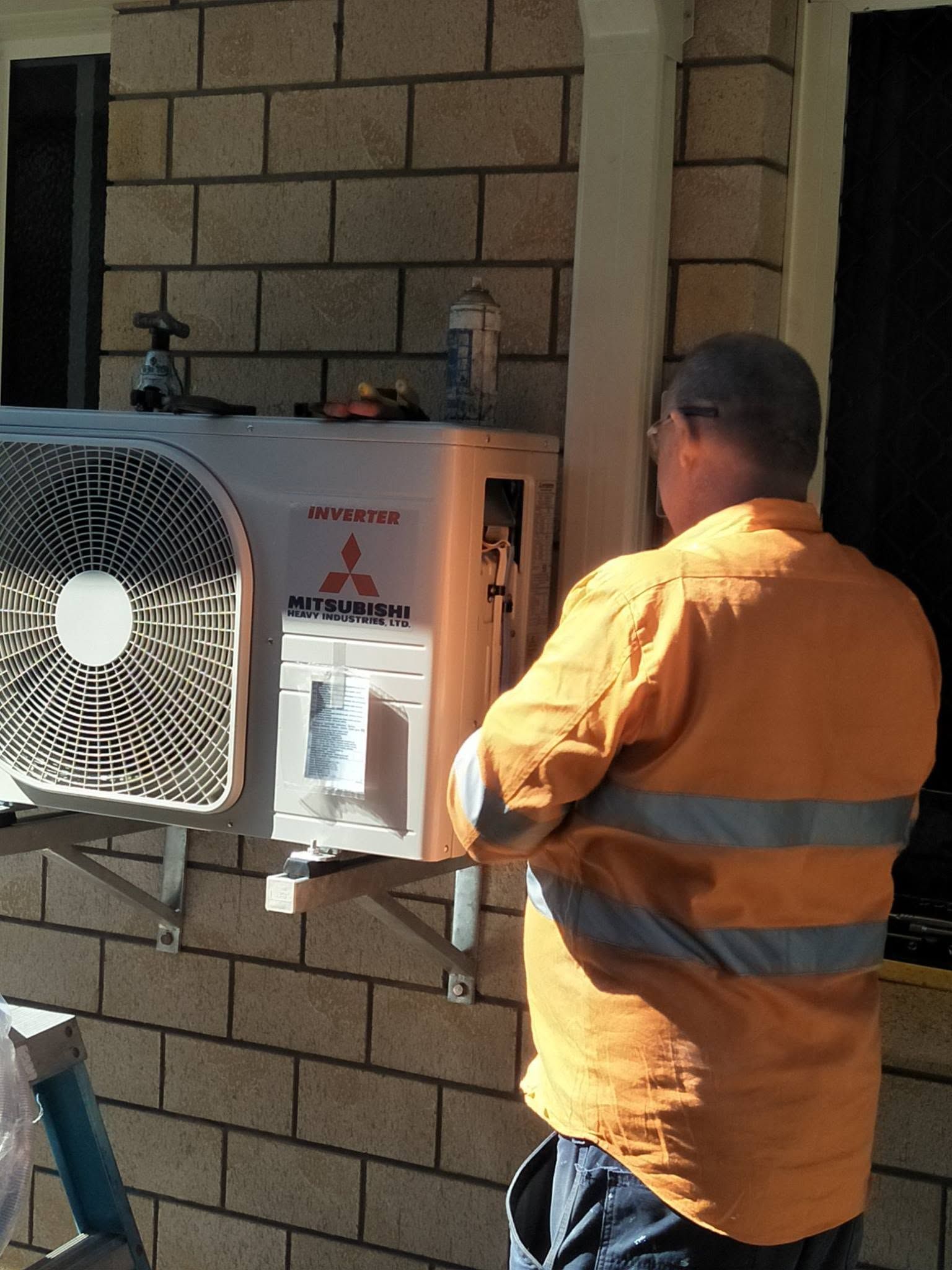 A Man In Orange Work Clothes Working On An Air Conditioning Unit Mounted On A Brick Wall — LSEA Electrical Group in Bucasia, QLD.