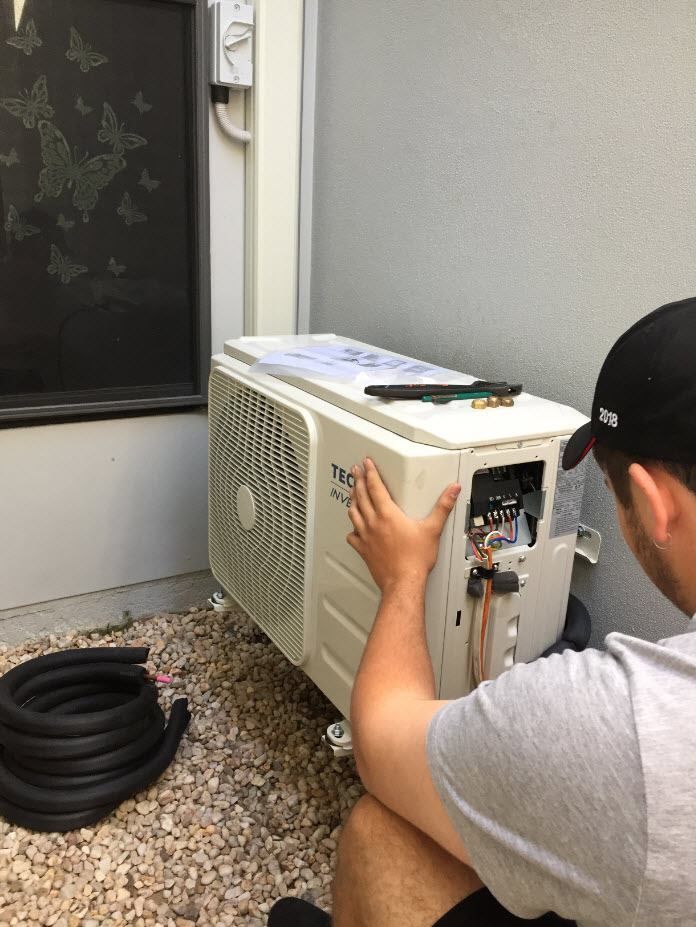 A Man Installs an Ac Unit Outside Against a Wall, With Copper Pipes and Tools Nearby — LSEA Electrical Group in Bucasia, QLD