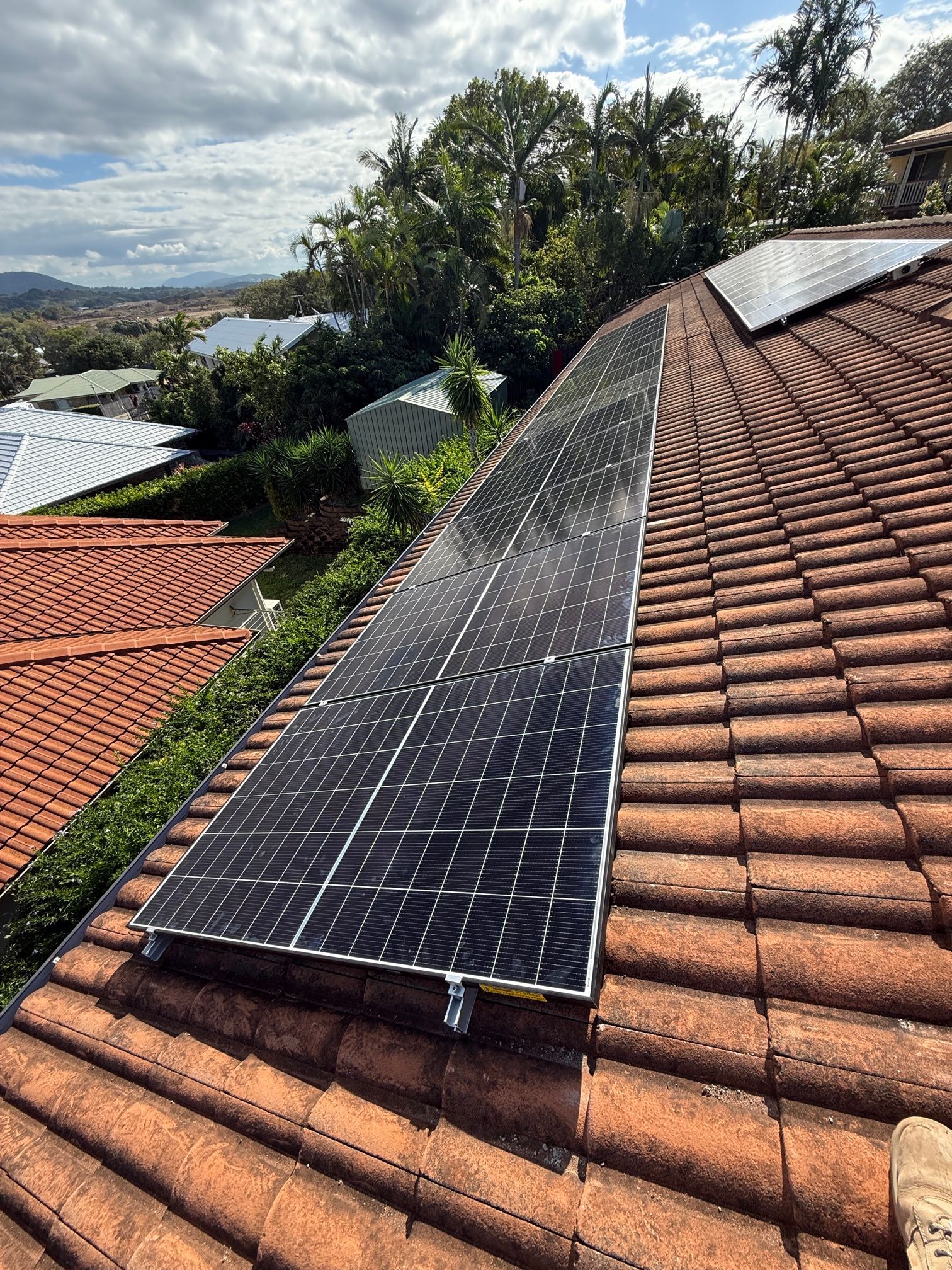 Solar panels mounted along a terracotta-tiled roof overlooking a hillside neighborhood under cloudy skies — LSEA Electrical Group in Bucasia, QLD