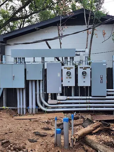 Row of gray electrical panels and conduit mounted on an exterior wall beneath a roof overhang.