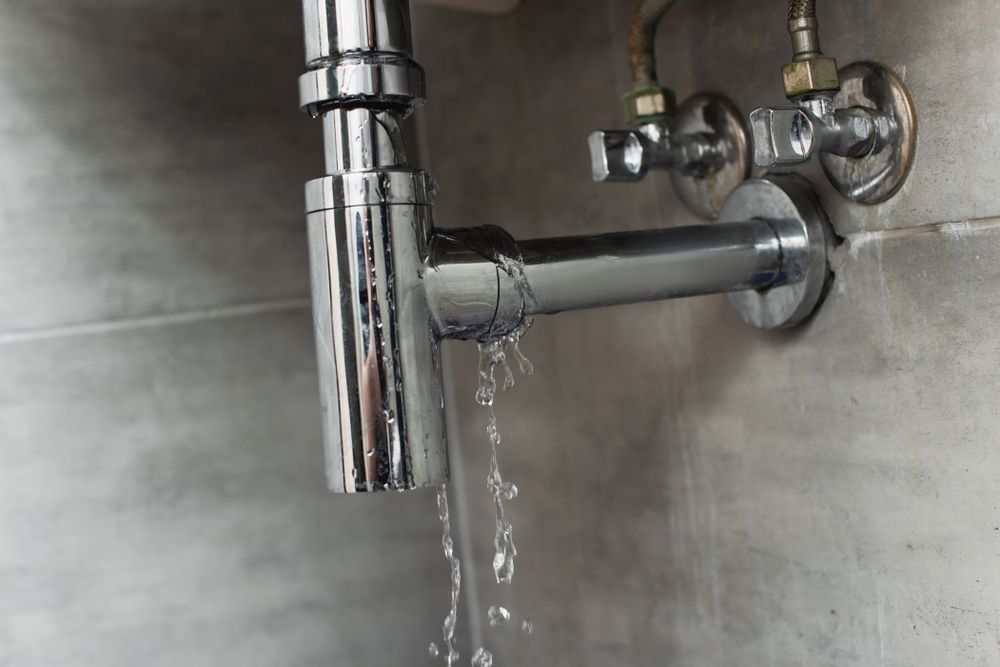 Leaking chrome sink pipe beneath a stainless steel counter, water dripping from the joint