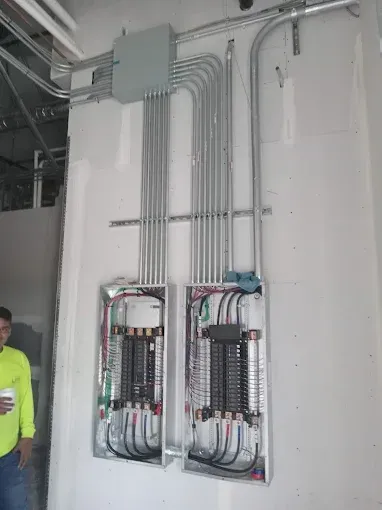 Electrical panel boxes with exposed wiring and conduit in a utility room; worker partly visible at left.