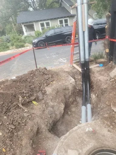 Utility trench with exposed pipes and a red caution tape barrier beside a street and parked cars