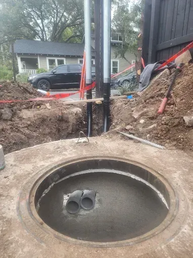 Open underground utility access hole at a construction site with exposed pipes and dirt excavation.