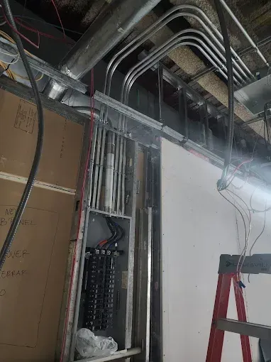 Construction site with exposed ceiling pipes, electrical wiring, and a red ladder against a wall.