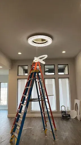 Two ladders under a ceiling light in a sparsely furnished room with windows and hardwood floors
