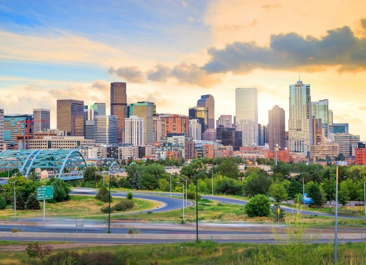 The Denver skyline at sunset with the Platte River Bridge in the foreground and a warm, cloudy sky above.