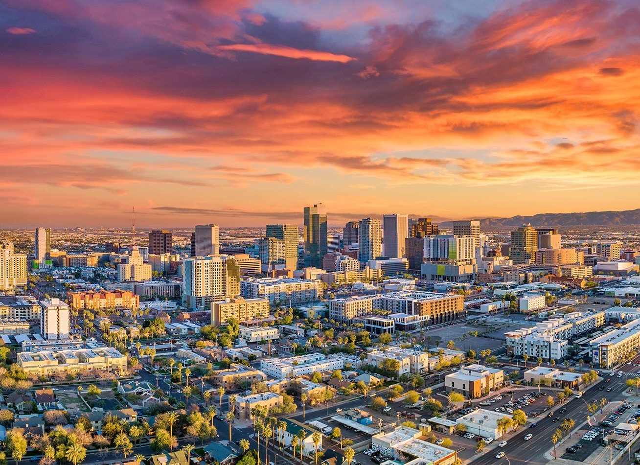 Aerial view of a city skyline at sunset with vibrant orange and pink clouds over high-rise buildings and suburban streets.
