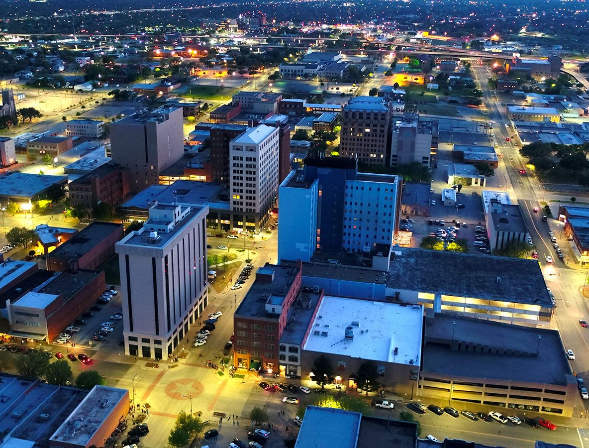 Aerial view of a city downtown at twilight, featuring illuminated skyscrapers, streets, and parking lots.