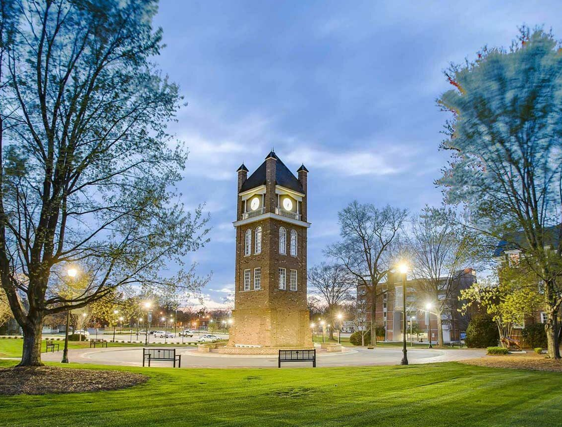 A lit brick clock tower stands in a grassy park at dusk, surrounded by trees and lamp posts.