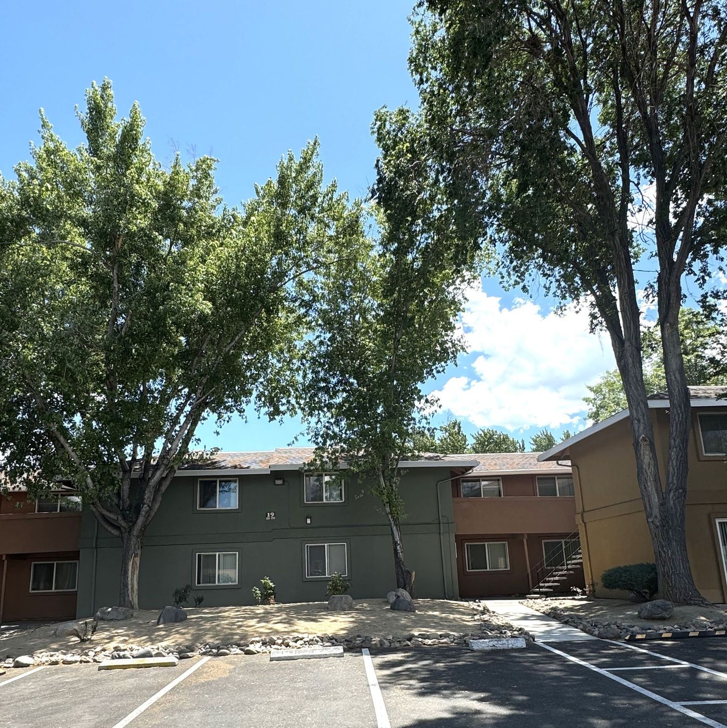 Apartment building exterior; green, brown, and tan paint, trees, blue sky, parking lot.