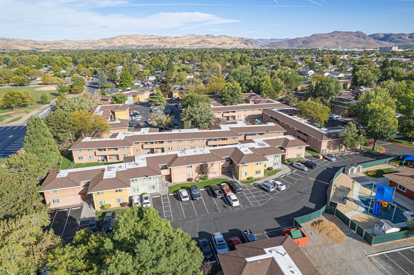 Aerial view of apartment complex with parking, playground, and mountain backdrop.