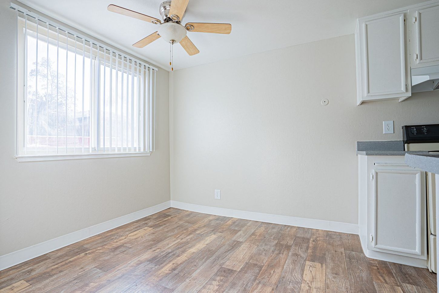 Empty room with wood-look floor, window with blinds, ceiling fan, and white kitchen cabinets.