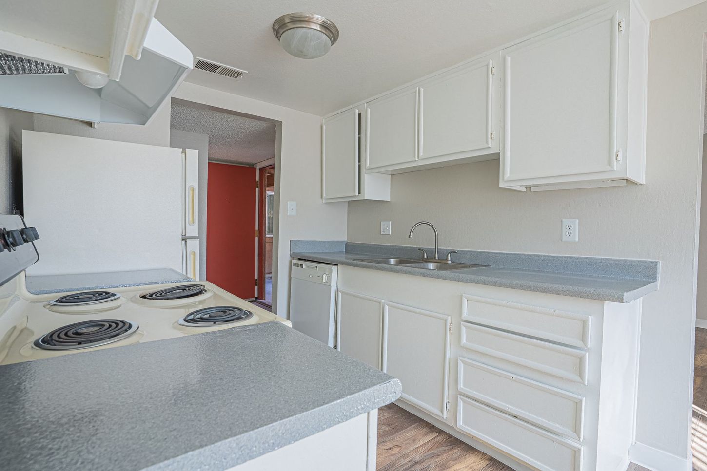 Kitchen with white cabinets, appliances, and gray countertop. A doorway with a red door is in the background.