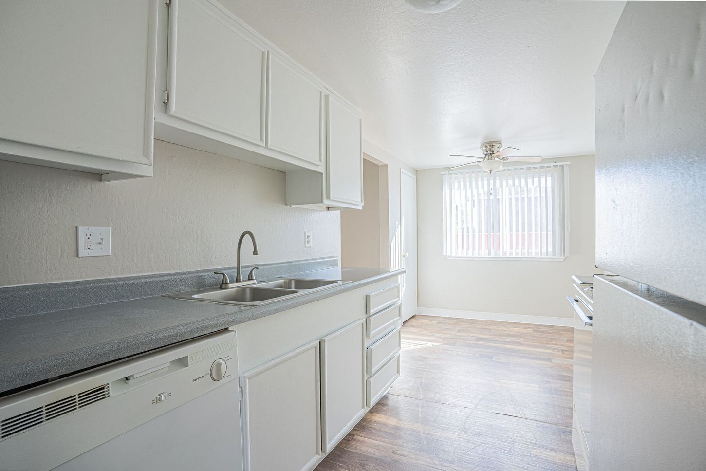White kitchen with cabinets, sink, and dishwasher. A window lets in light, visible hardwood floor.