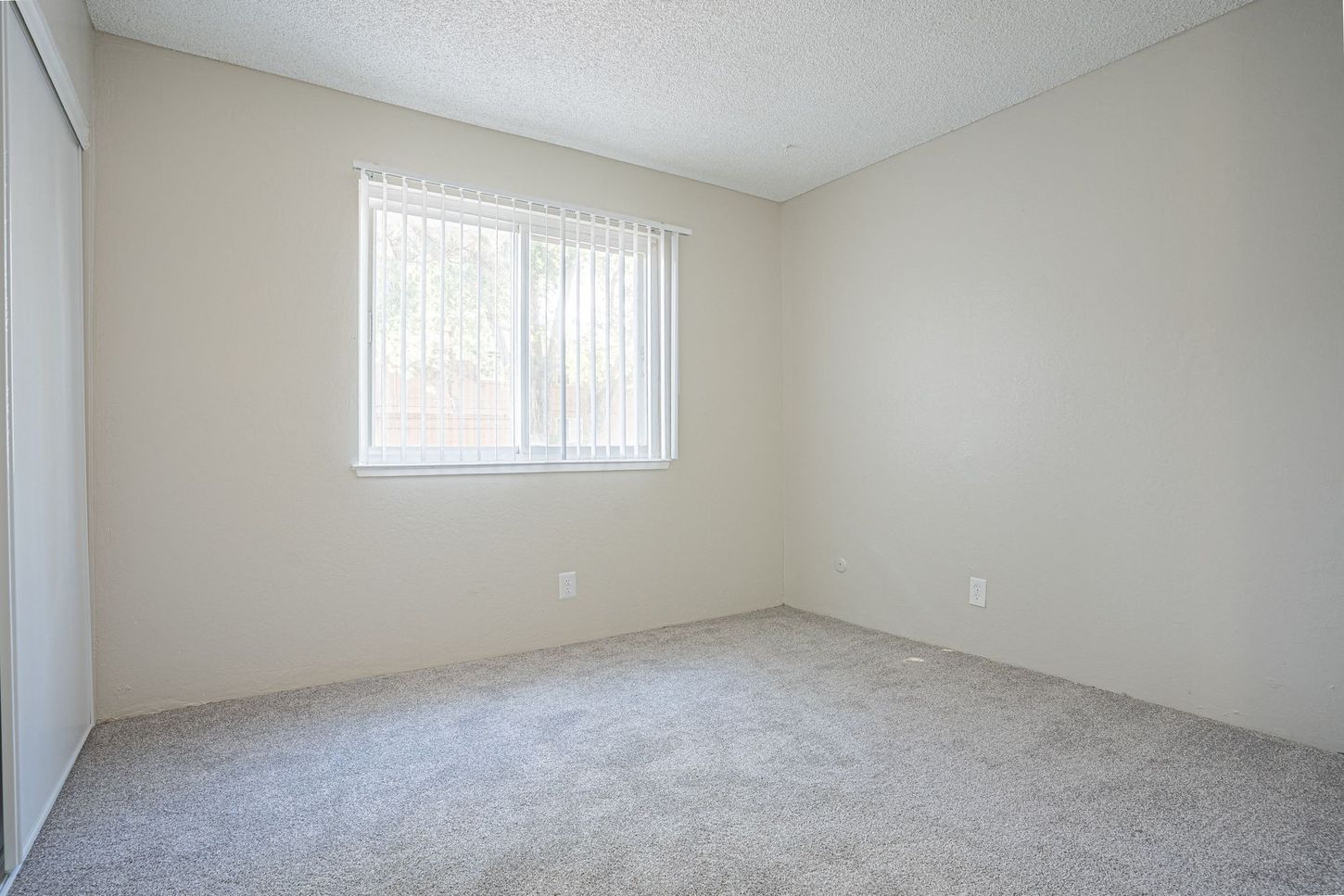 Empty room with light gray carpet, walls, and ceiling; a window with sheer curtains.