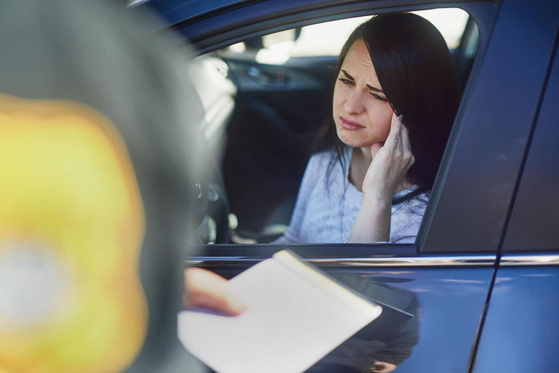 woman sitting inside a blue car, interacting with a person outside the vehicle holding a notepad.