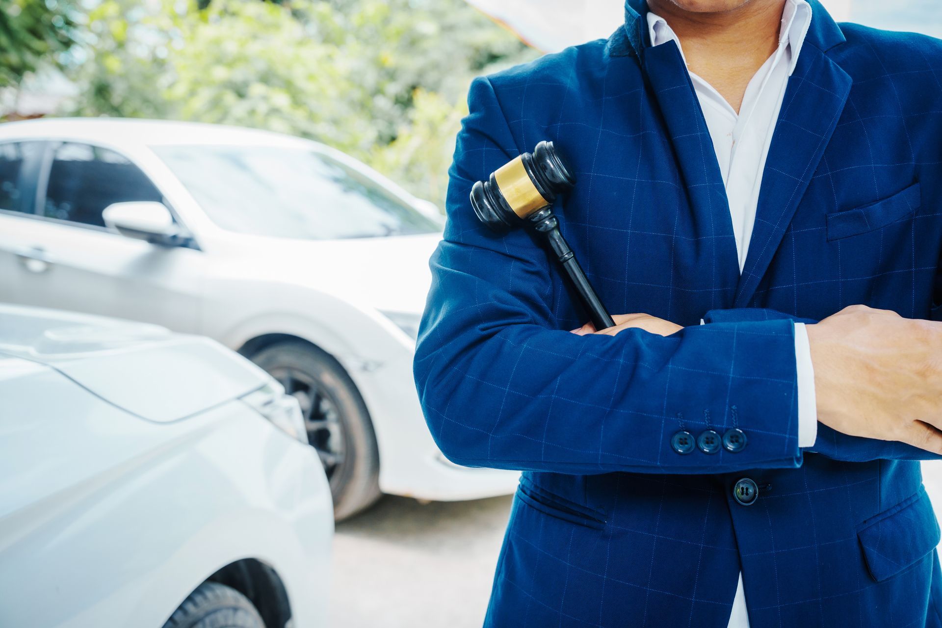 A DUI lawyer with folded arms, holding a wooden gavel, stands next to a car with another car in the 