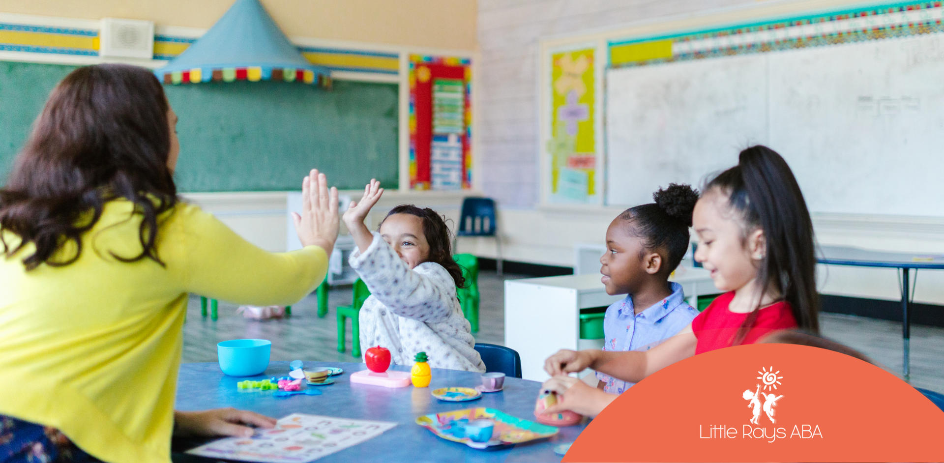 Teacher gives high-five to a student in a classroom with other children; blue table, green chairs.