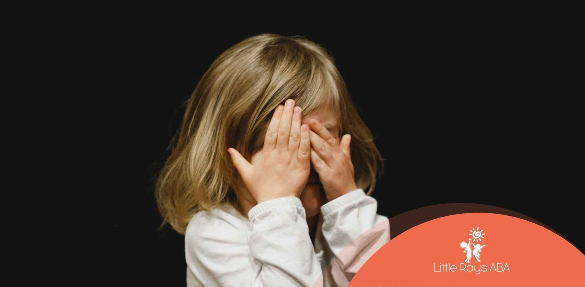 A child with blonde hair covering their eyes with their hands, expressing fear or shyness.