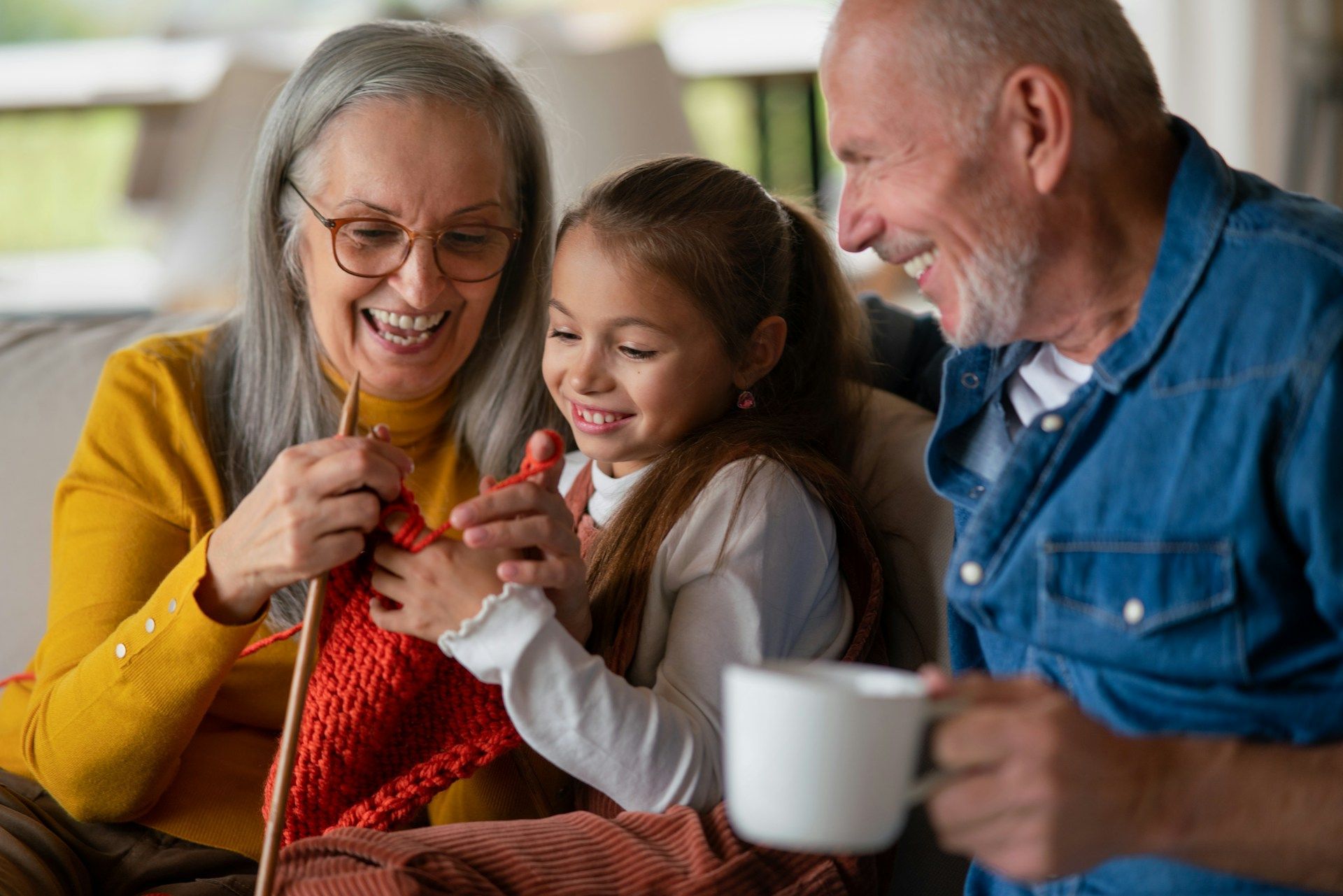 An elderly couple and a little girl are knitting together while sitting on a couch.