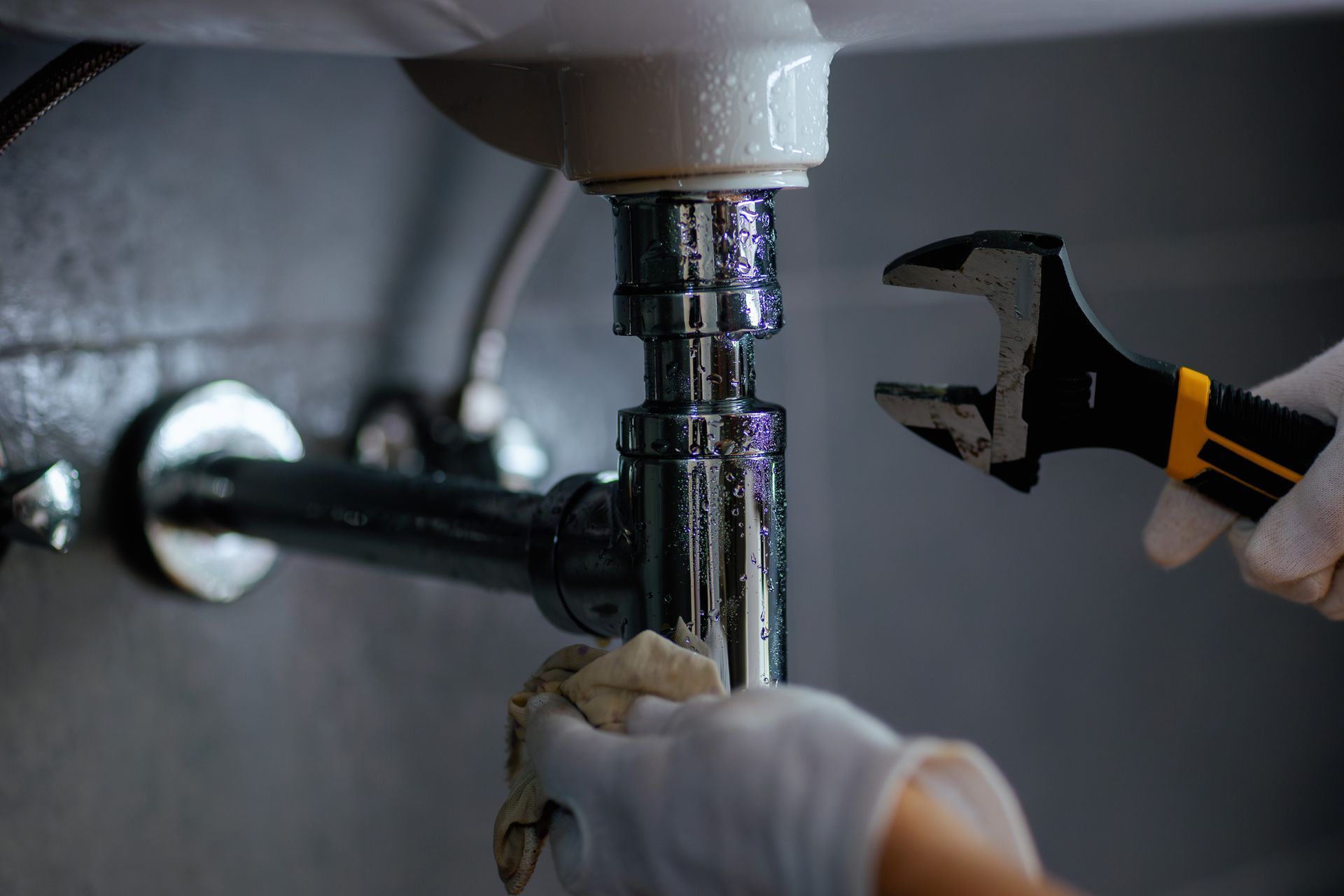 Hands with gloves, fixing a leaky sink with a wrench and cloth.
