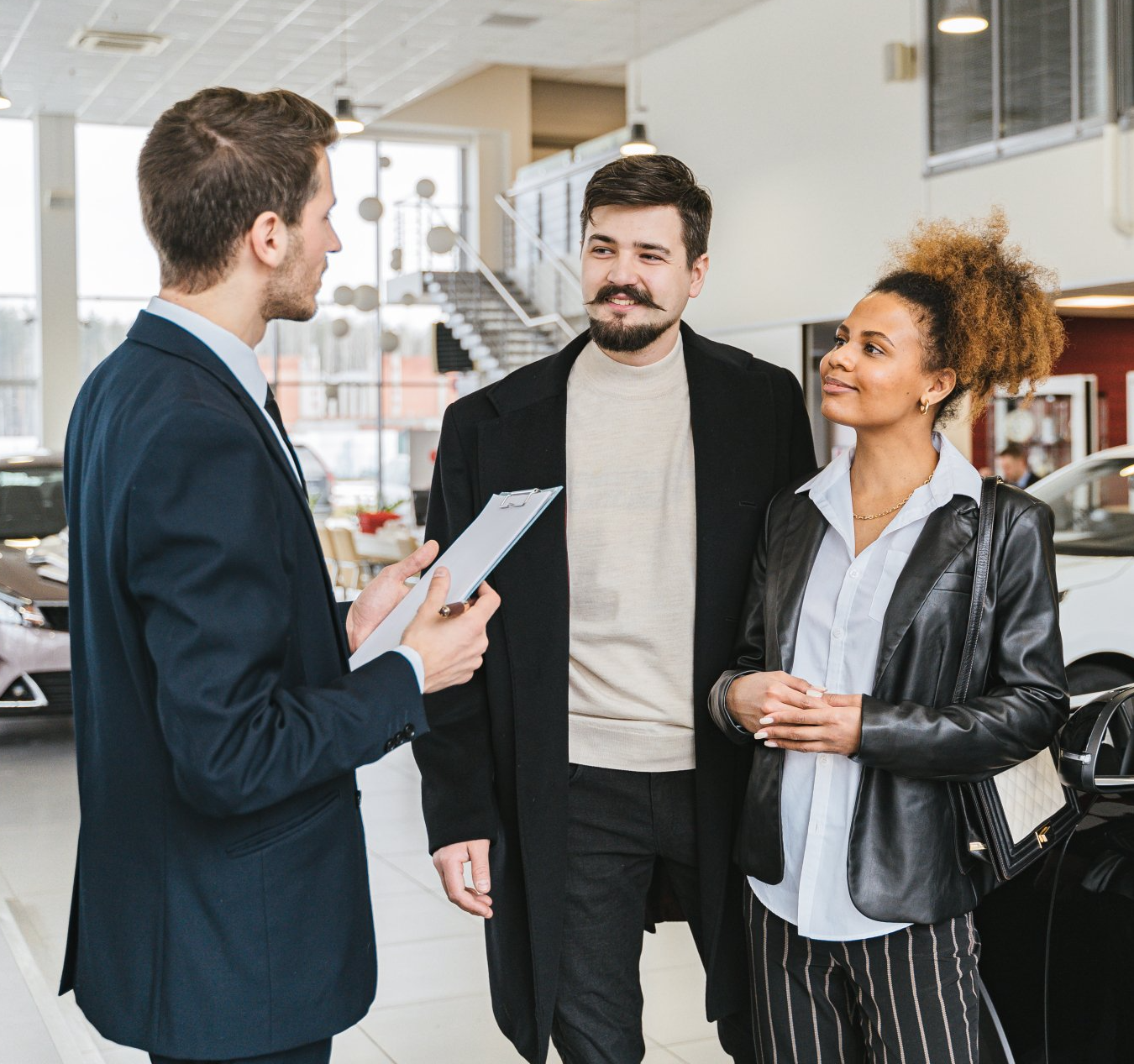 A man in a suit is talking to two people in a car showroom.