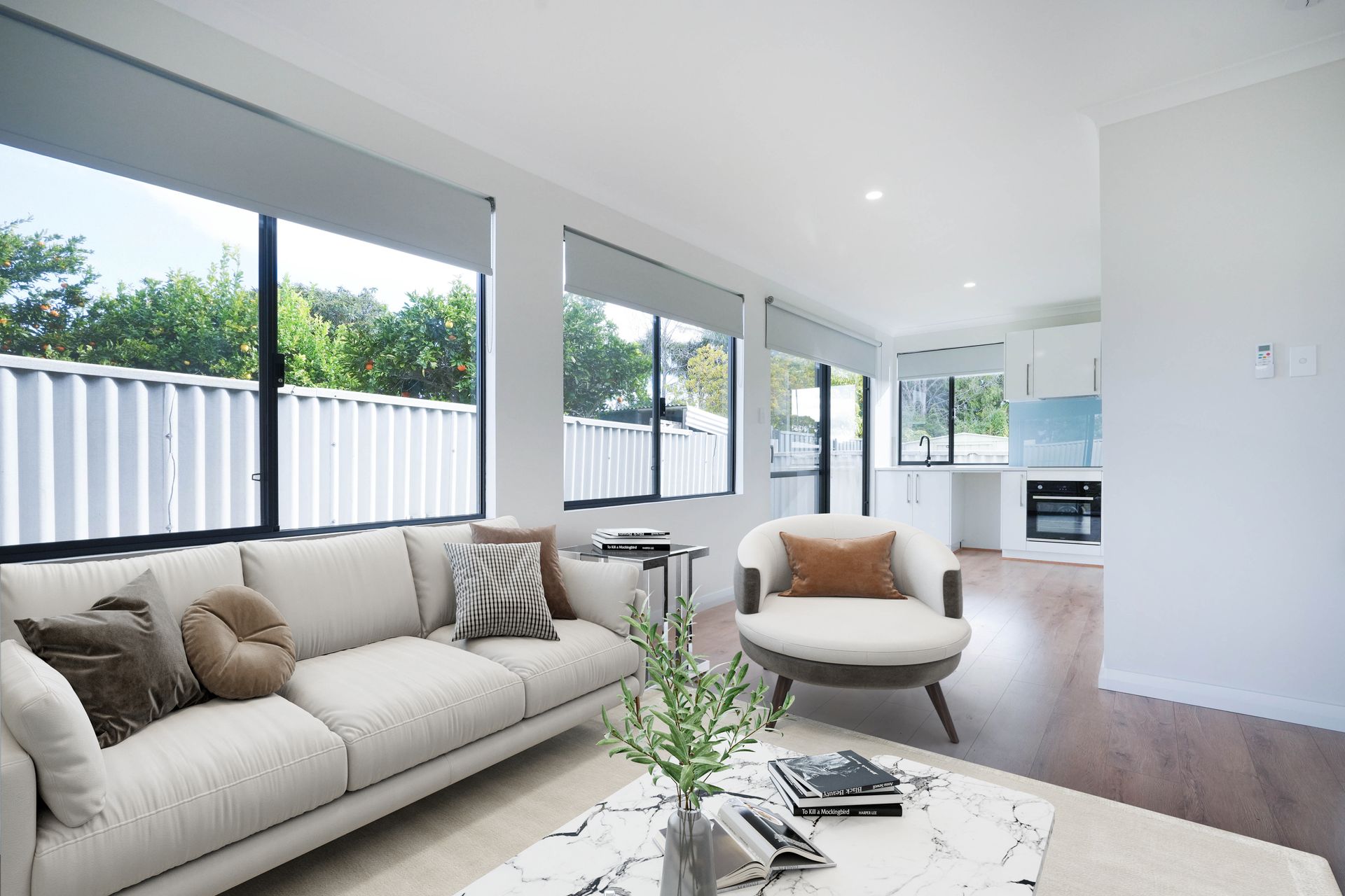 A bright, modern living room with a beige sofa, a round armchair, and large windows overlooking a backyard fence.