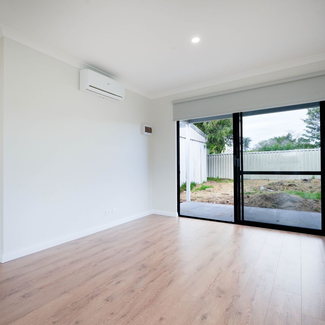 A bright, empty room with light wood-look flooring, white walls, a mounted air conditioner, and a large sliding glass door.