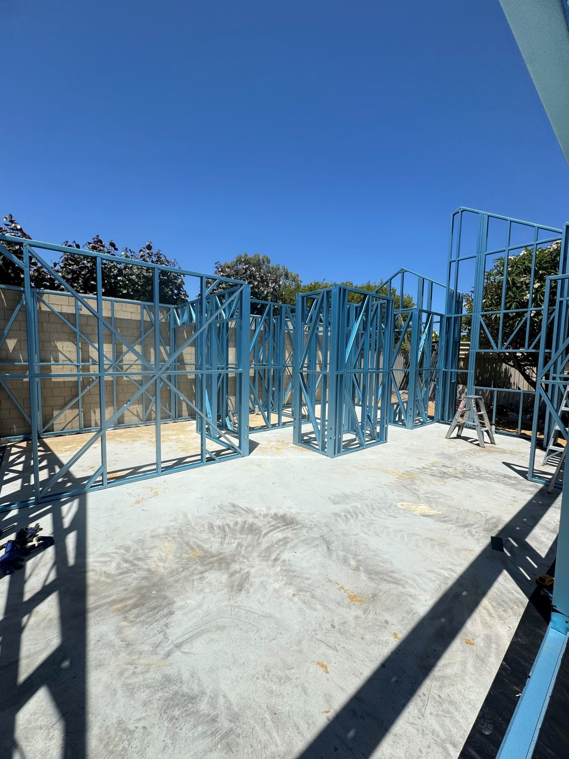 A construction site showing light-gauge steel wall framing erected on a concrete foundation under a clear blue sky.