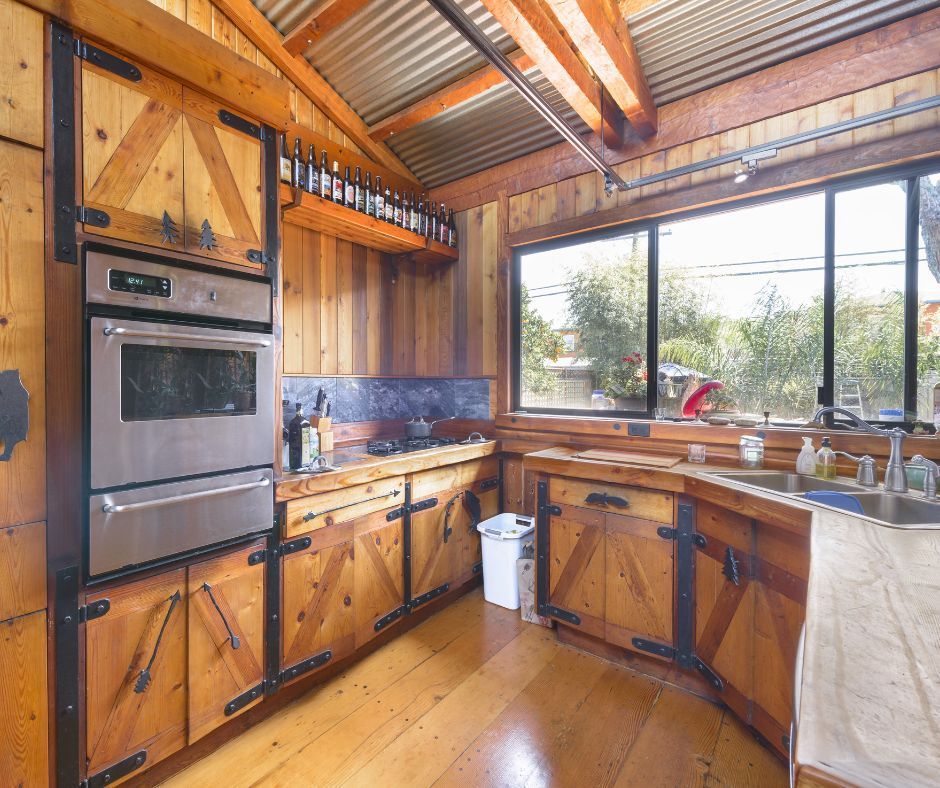 Rustic kitchen with wood-paneled walls and cabinets, stainless steel oven, corrugated metal ceiling, and large windows.