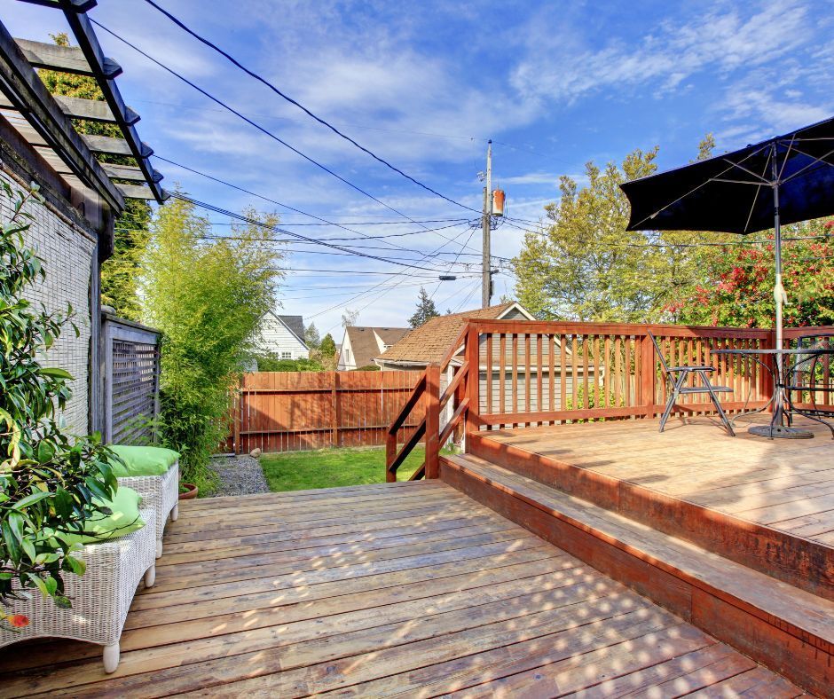 A sunny wooden deck with a black patio umbrella, garden furniture, and a view of a backyard with a wooden fence.