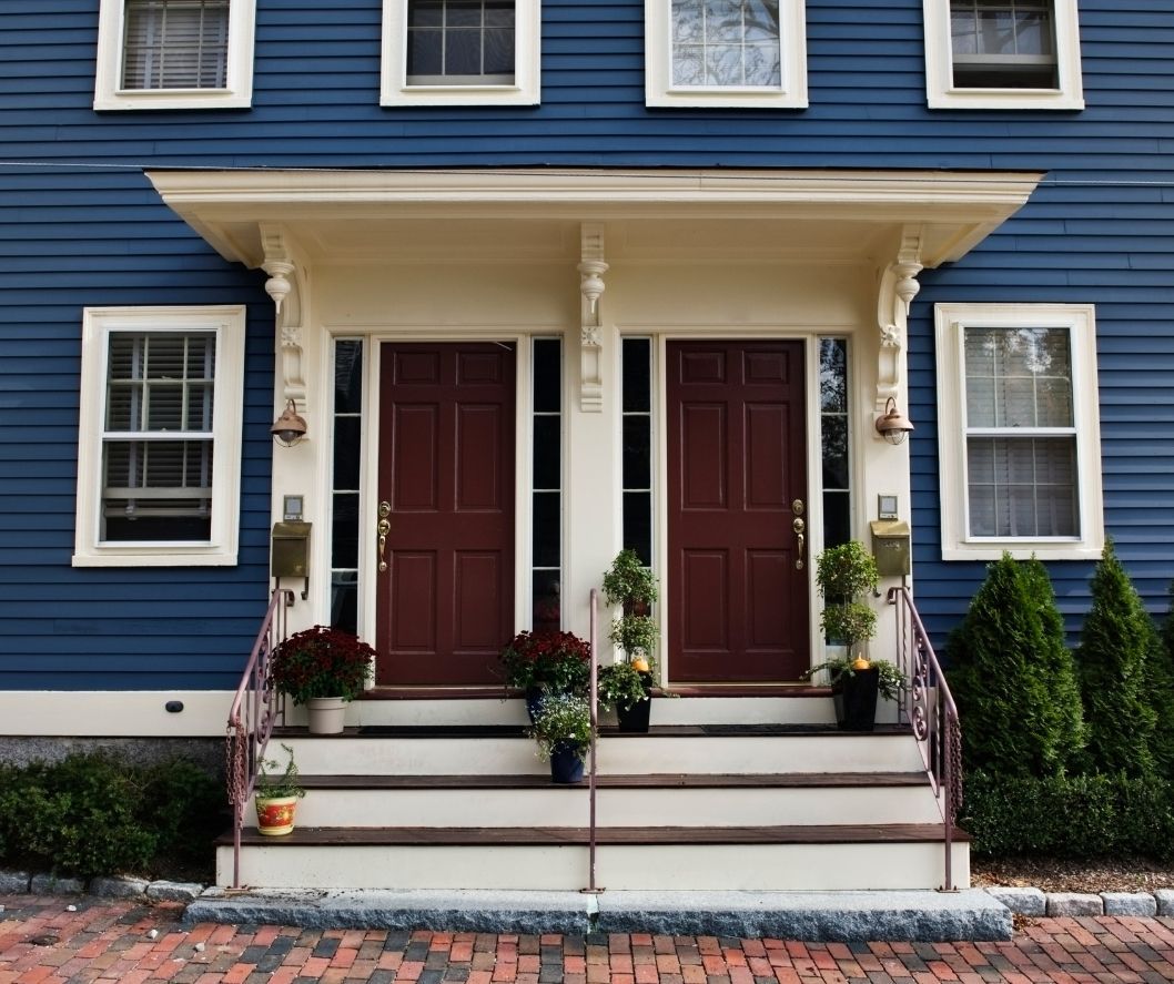 Two dark red front doors side-by-side on a blue house, featuring a shared porch roof, white trim, and stone steps.