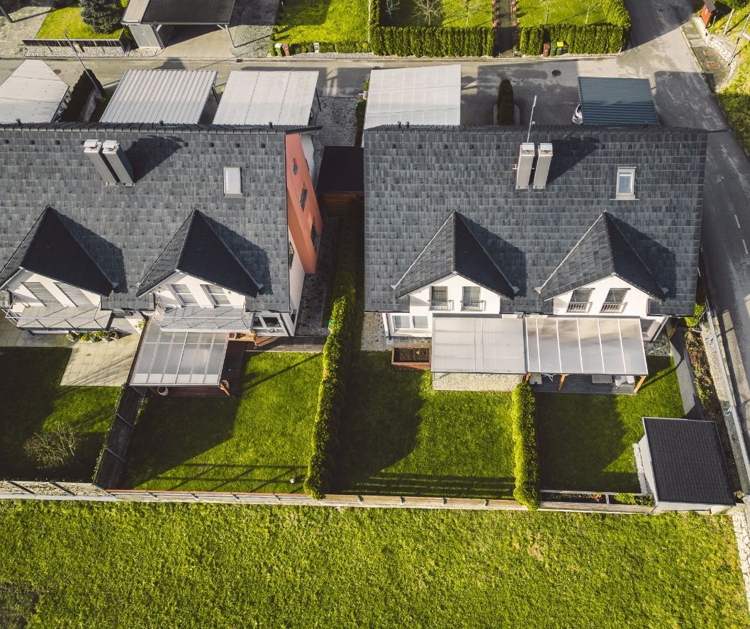 An aerial view of two rows of houses with dark gray roofs, green backyards, and privacy hedges.