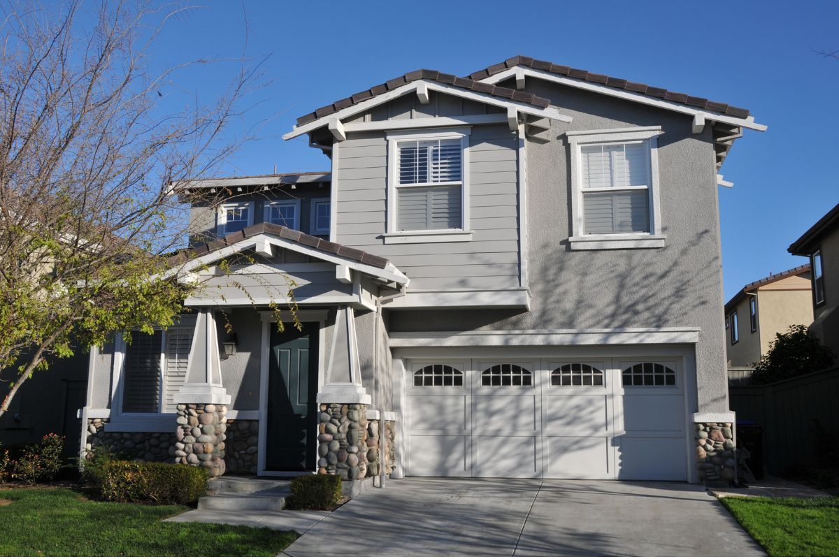 A two-story grey suburban house with stone accents on the front porch and a three-car garage under a clear blue sky.