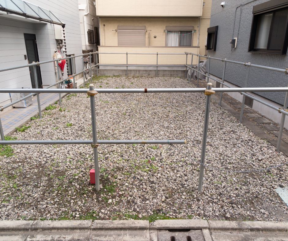 A vacant rectangular lot covered in gravel, enclosed by a simple metal pipe fence, situated between residential buildings.