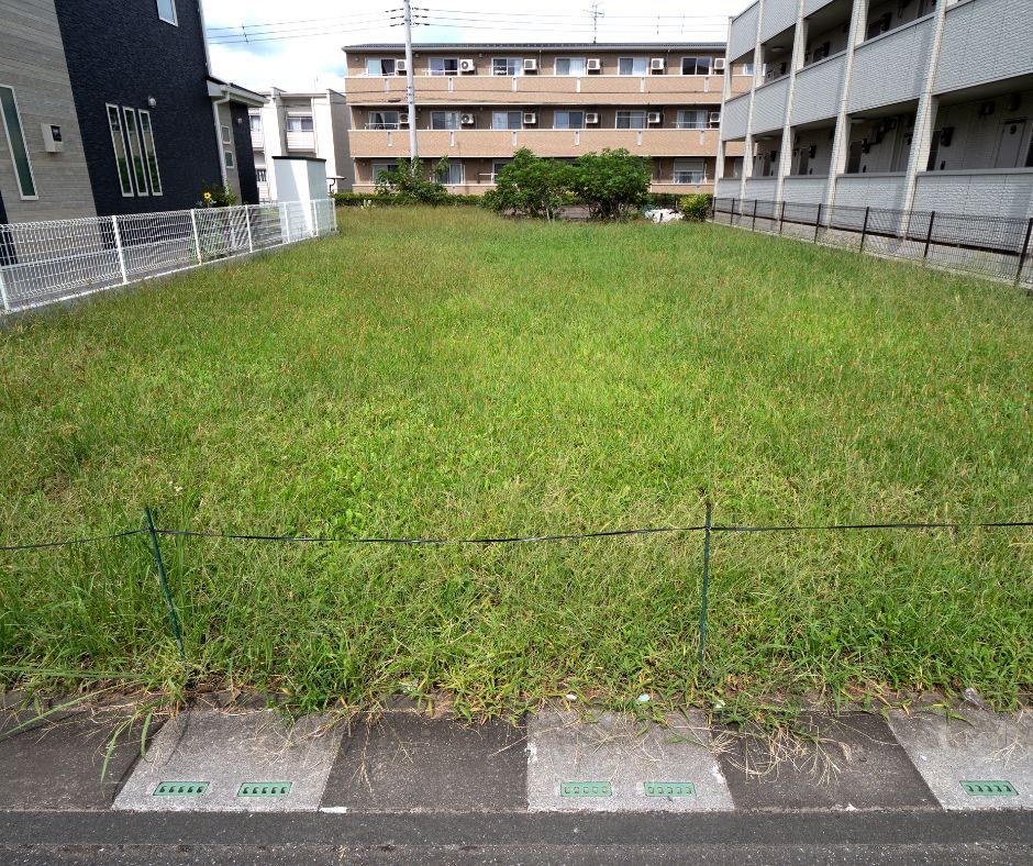 A rectangular, grass-covered vacant lot sits between a residential building and an apartment complex.