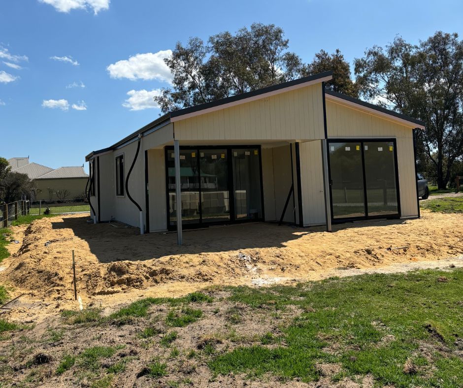 A new single-story house with cream siding and black-framed sliding glass doors, surrounded by sandy, unfinished landscaping.