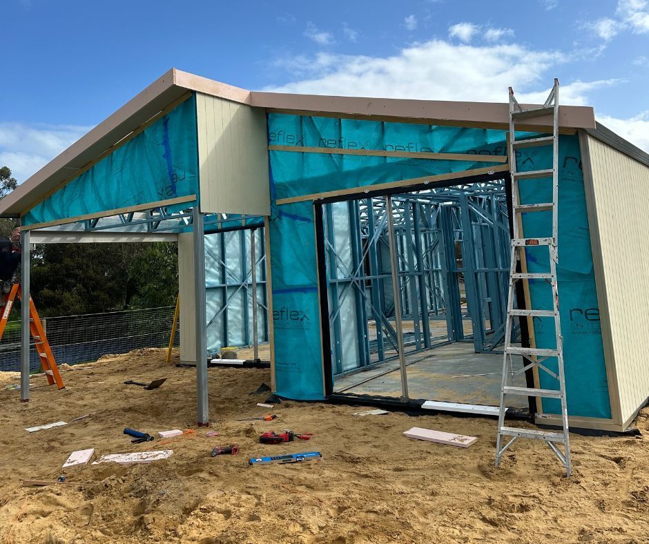 A steel-framed building under construction with blue protective wrap, standing on a sandy site with ladders nearby.