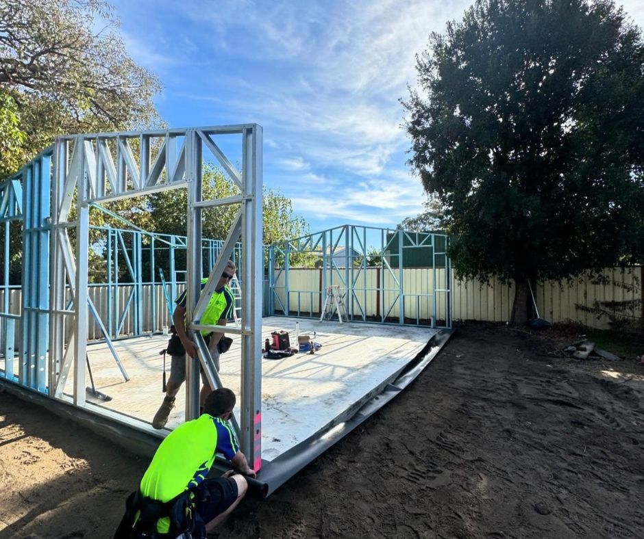Two construction workers in high-visibility vests assemble a steel frame for a residential structure outdoors.