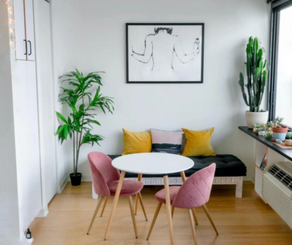 A small dining area with two pink velvet chairs, a round white table, a bench with mustard pillows, and potted plants.