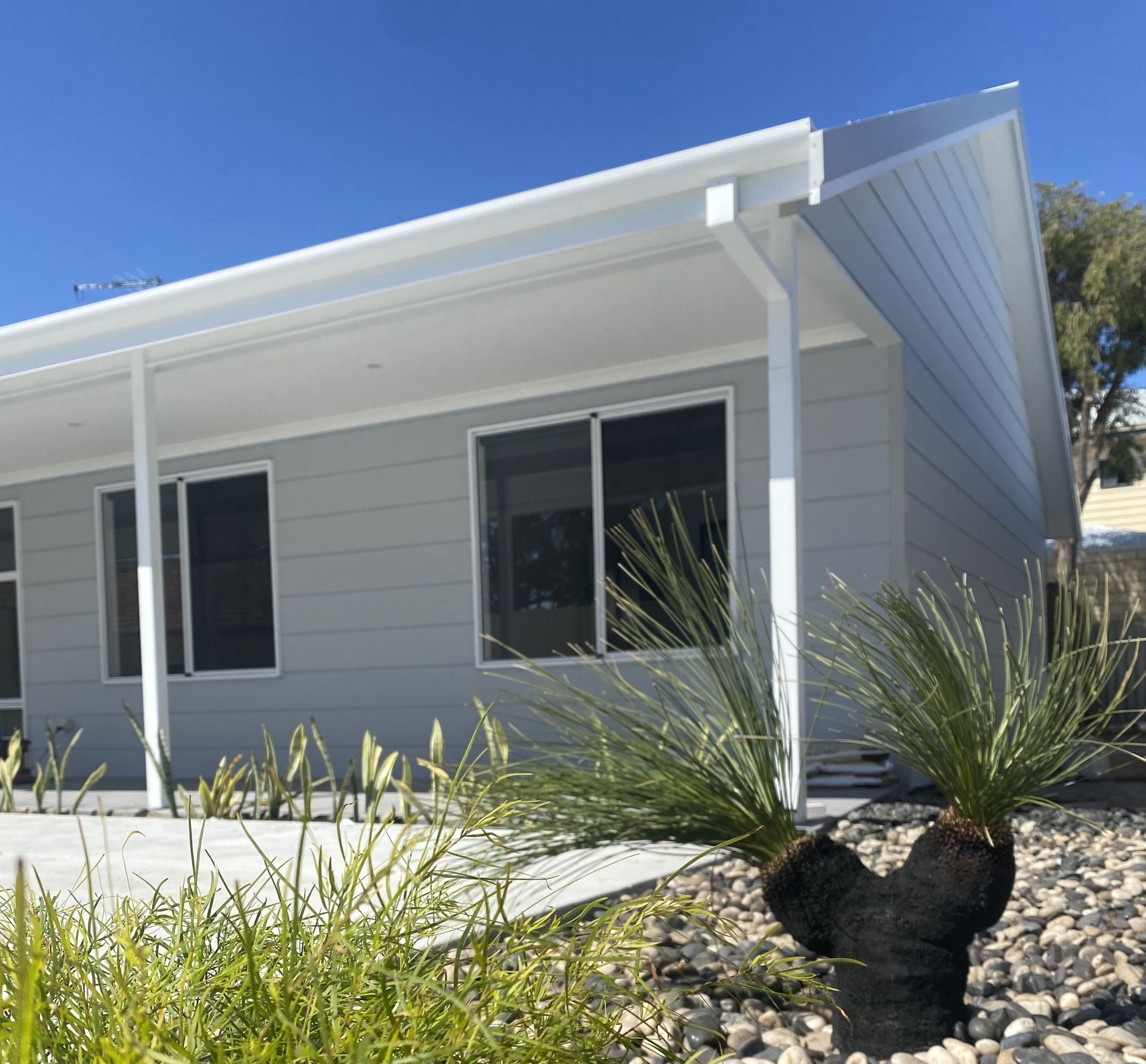 A light gray modern house with a white-pillared porch, a rock garden, and a distinctive grass tree under a blue sky.